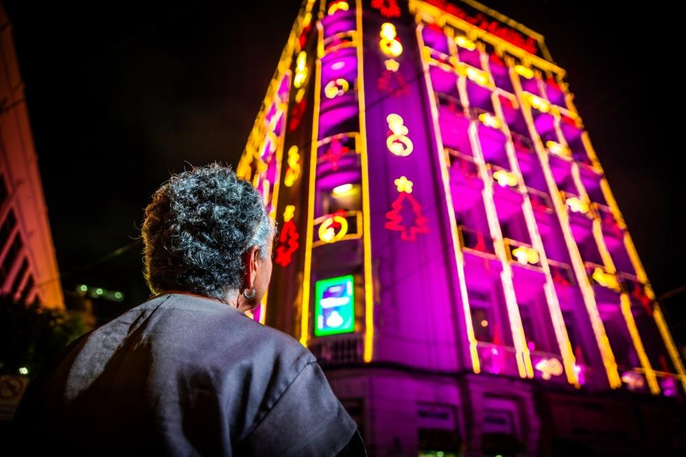 Na imagem, uma foto noturna tirada em ângulo de contraponto (de baixo para cima), focando nas costas e no ombro direito de uma mulher com cabelo curto e grisalho. Ela usa uma blusa escura de manga curta e um brinco pequeno e prateado. A mulher está olhando para um prédio alto, que domina o centro da imagem. O prédio está intensamente decorado com luzes de Natal. A decoração é predominantemente neon/LED nas cores magenta ou roxo vibrante, com contornos amarelos/dourados, e inclui formas de estrelas e árvores de Natal vermelhas/rosas brilhantes. O céu noturno é completamente escuro, e a iluminação colorida do prédio contrasta fortemente com o fundo.