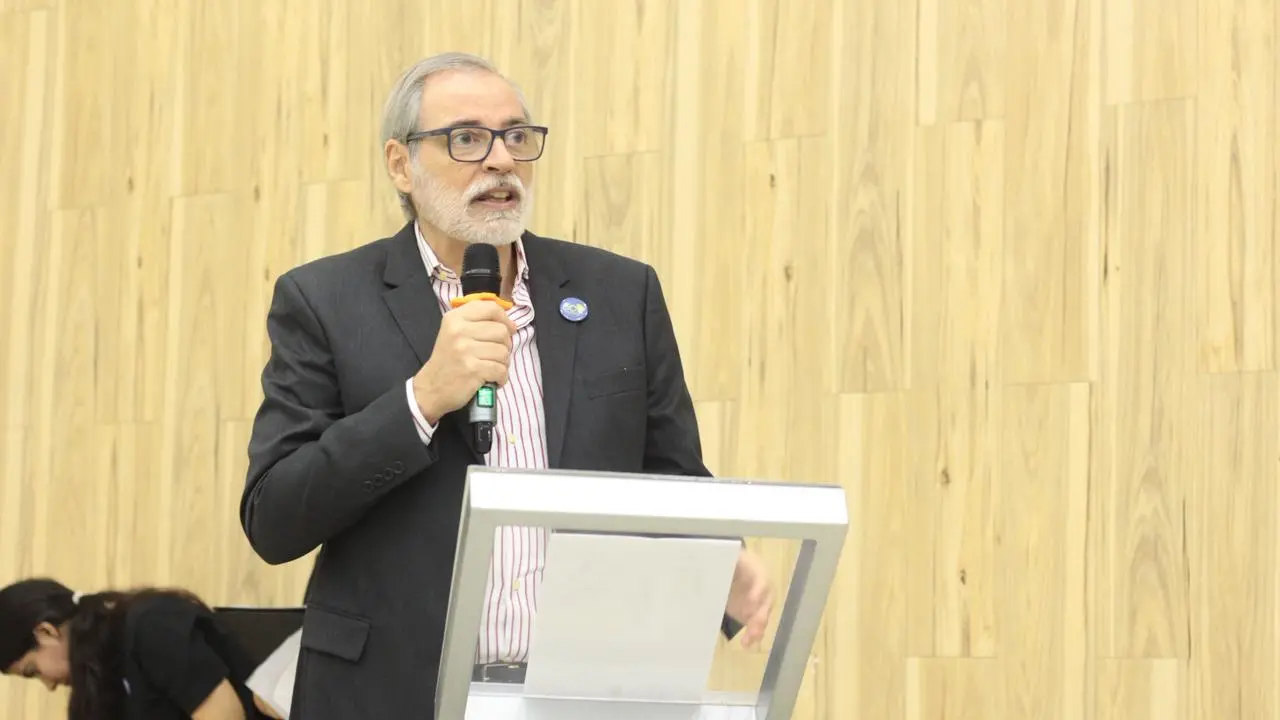 Homem de pele clara, com cabelos grisalhos e barba curta também grisalha, fala ao microfone em um púlpito. Ele usa óculos de armação escura, um paletó cinza-chumbo sobre uma camisa social listrada de rosa e branco, e exibe um pequeno broche azul na lapela. Ele segura o microfone com a mão direita e parece estar em meio a um discurso. Ao fundo, uma parede revestida com painéis de madeira clara em tons verticais. No canto inferior esquerdo, parte da cabeça de uma pessoa com cabelos escuros é visível.