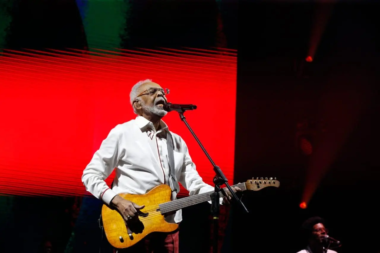 Cantor Gilberto Gil se apresenta no palco com violão, braços abertos diante do público, durante show com telão ao fundo.