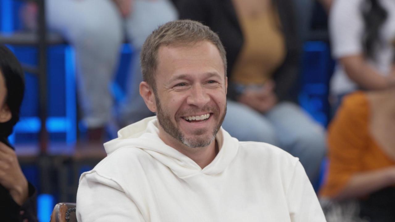 Tiago Leifert com moletom branco, sorrindo e olhando para o lado, de frente para uma plateia.