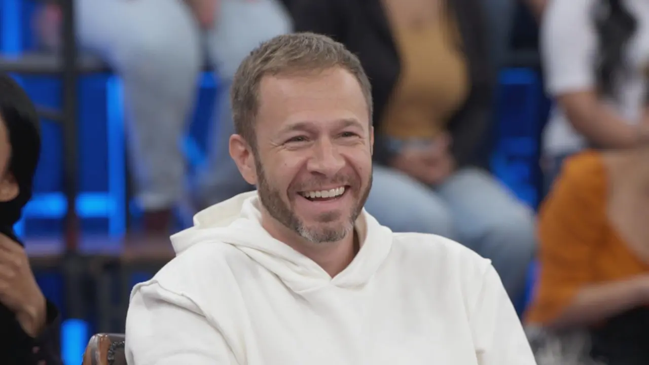 Tiago Leifert com moletom branco, sorrindo e olhando para o lado, de frente para uma plateia.