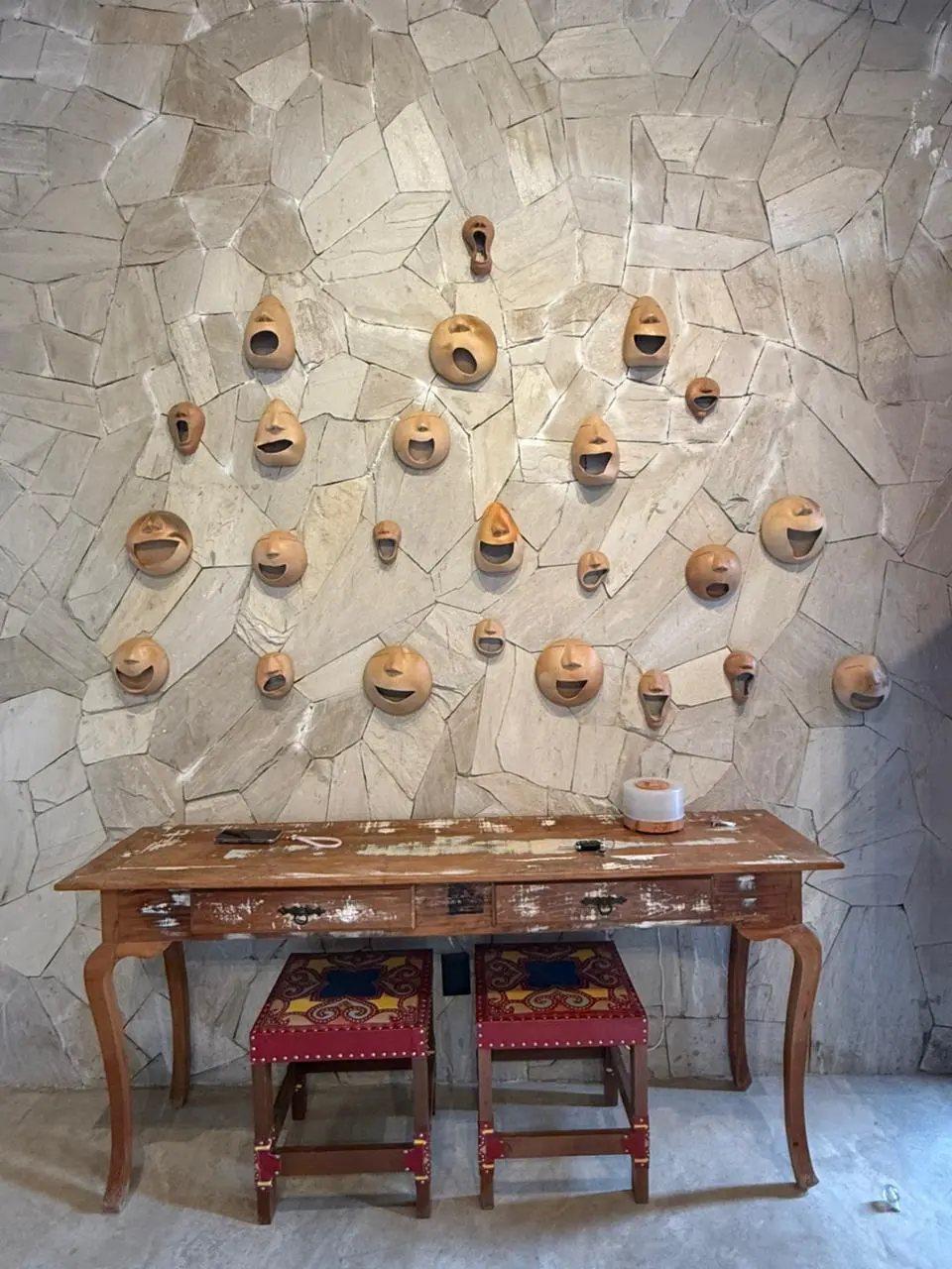 Na imagem, close-up de uma parede de destaque revestida com pedras irregulares de tom bege claro. Pendurada na parede, há uma coleção de máscaras ou cabeças de cerâmica em tons de terracota, cada uma com expressões faciais variadas, como sorrisos e bocas abertas. Abaixo da composição, encontra-se um aparador de madeira rústica com acabamento desgastado. Sob o aparador, estão guardados dois bancos (pufes) quadrados com estofamento vermelho e padrões coloridos de inspiração folclórica.