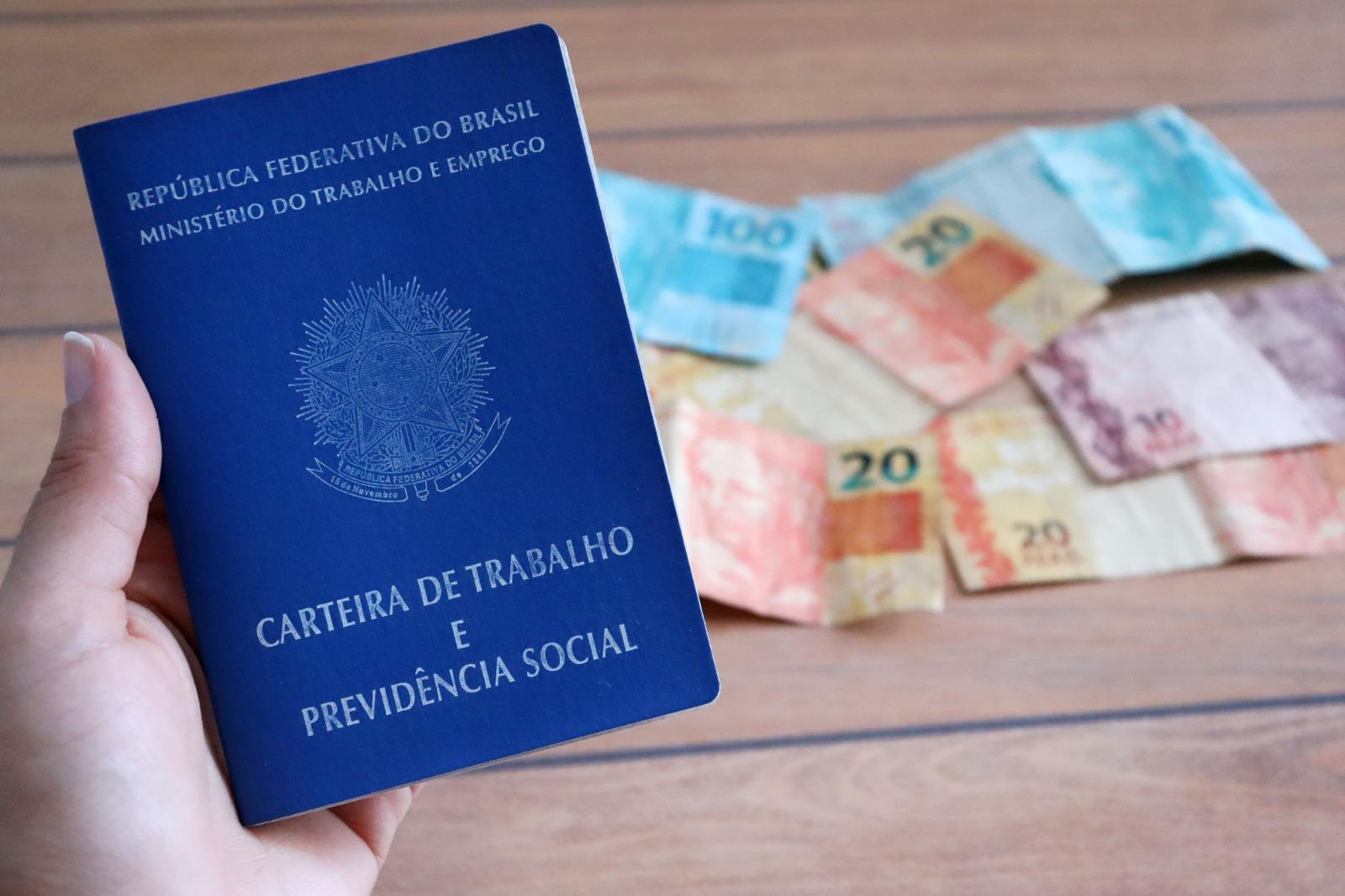 Uma pessoa segura uma carteira de trabalho com uma quantia em dinheiro atrás.