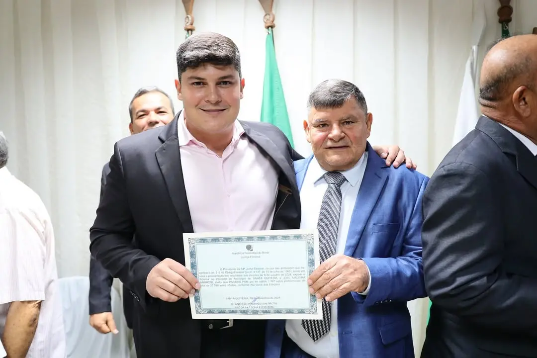 Dois homens posam lado a lado em um ambiente formal, vestindo ternos. Eles seguram juntos um certificado com moldura clara, exibido em frente ao corpo. O homem à esquerda usa camisa clara e sorri levemente; o homem à direita usa terno azul e gravata, com expressão séria. Ao fundo, há cortinas claras e bandeiras em tons de verde e branco, além de outras pessoas parcialmente visíveis, sugerindo uma cerimônia oficial.