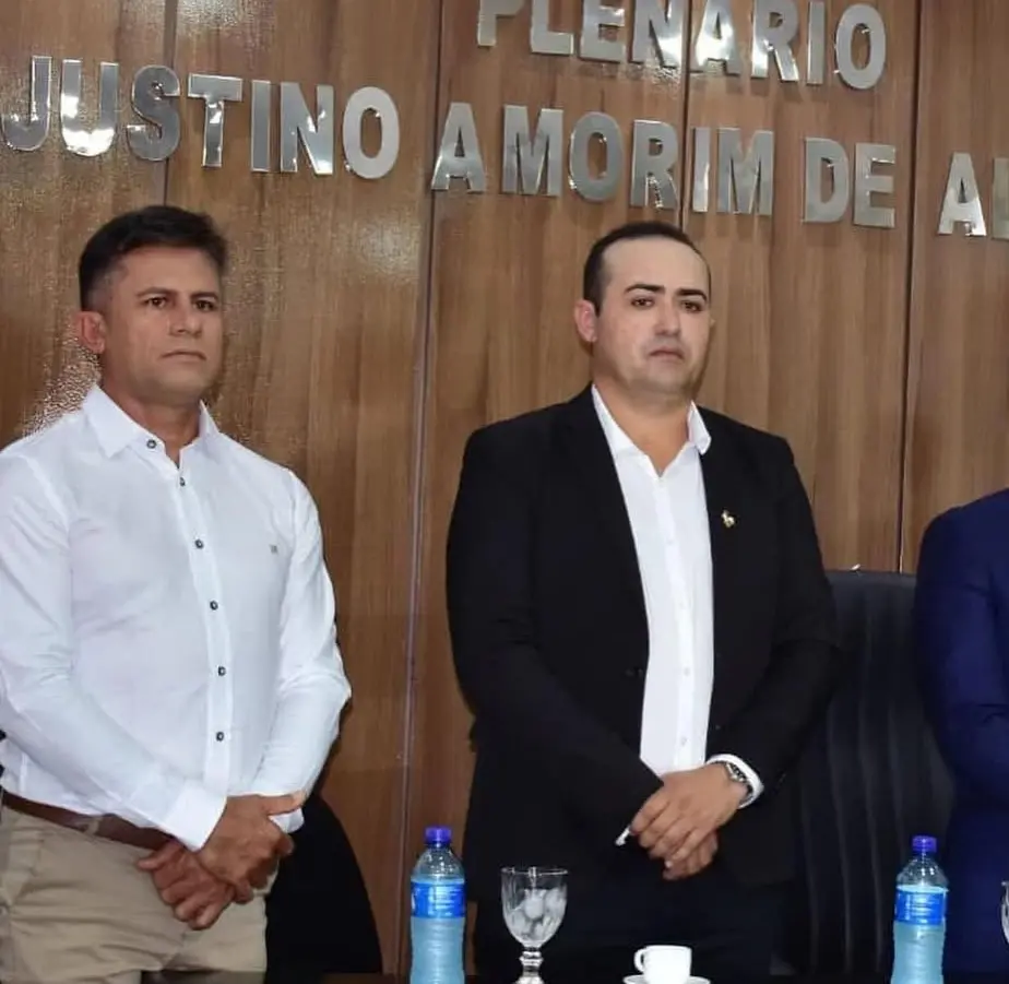 Dois homens estão em pé, lado a lado, em um ambiente institucional, diante de uma parede de madeira com letras metálicas que indicam o nome de um plenário. Ambos vestem roupas formais: o homem à esquerda usa camisa branca e calça clara; o homem à direita usa camisa branca e paletó escuro. Eles mantêm postura séria, com as mãos à frente do corpo. À frente, sobre uma mesa, há garrafas de água, copos e uma xícara, sugerindo uma sessão ou cerimônia oficial.