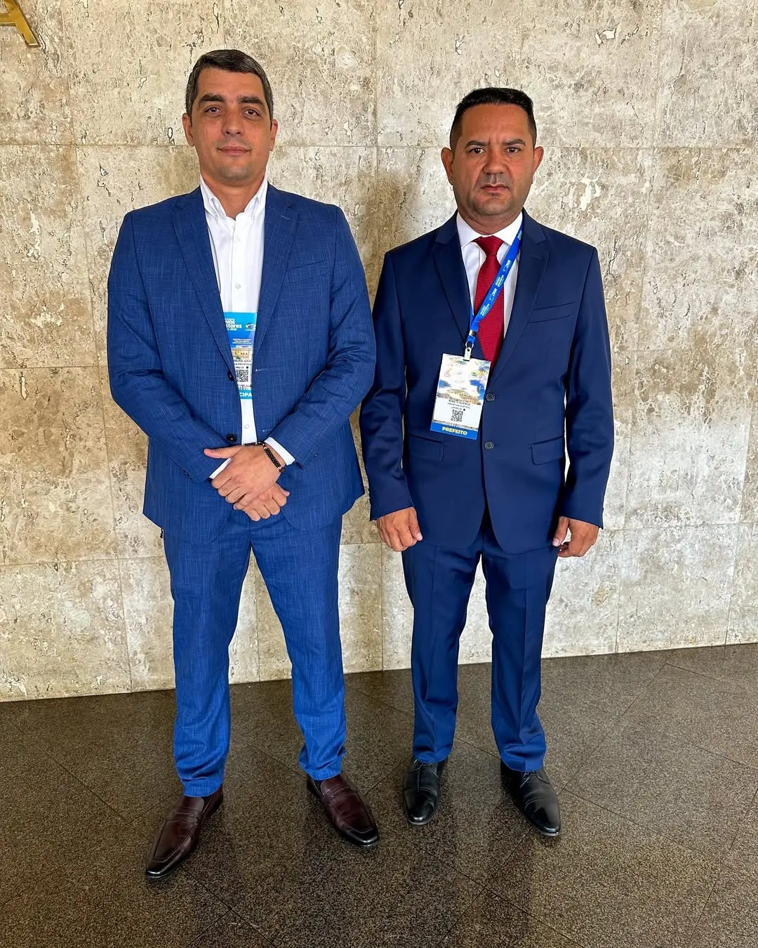 Dois homens posam em pé, lado a lado, em um ambiente interno com parede de pedra clara ao fundo. Ambos vestem ternos azuis e camisas claras; o homem à esquerda está sem gravata, com as mãos cruzadas à frente do corpo, e o homem à direita usa gravata vermelha e um crachá pendurado no pescoço. Eles olham para a câmera com expressão séria. O chão é escuro e polido, sugerindo um espaço institucional ou de evento oficial.