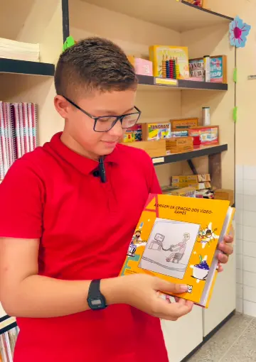 Foto do menino Isaac, estudante do quinto ano da escola Municipal Naiza Lira Rocha, ao lado do livro que escreveu e foi publicado pelo colégio.
