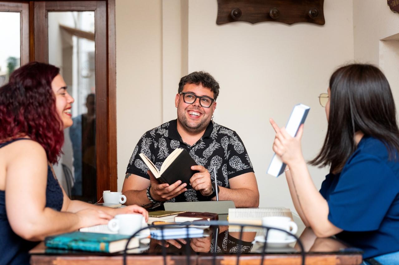 Fotografia de três jovens adultos sentados ao redor de uma mesa de vidro em um ambiente interno iluminado. No centro, um homem sorridente de óculos e camisa preta estampada segura um livro aberto. À esquerda, uma mulher de perfil com cabelos avermelhados sorri, e à direita, outra mulher de costas com blusa azul gesticula enquanto segura um livro. Sobre a mesa, há diversas xícaras de café e livros espalhados.
