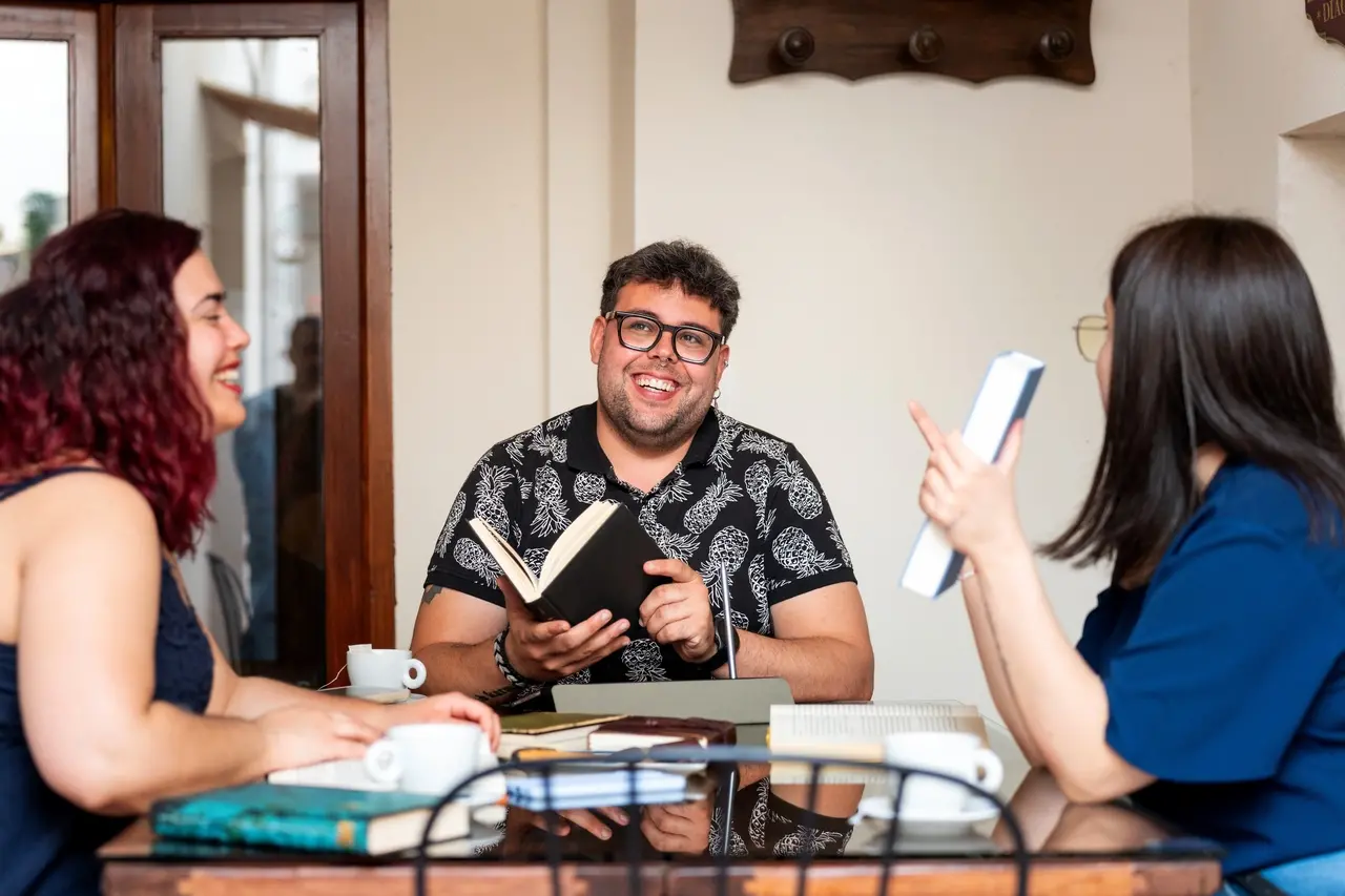 Fotografia de três jovens adultos sentados ao redor de uma mesa de vidro em um ambiente interno iluminado. No centro, um homem sorridente de óculos e camisa preta estampada segura um livro aberto. À esquerda, uma mulher de perfil com cabelos avermelhados sorri, e à direita, outra mulher de costas com blusa azul gesticula enquanto segura um livro. Sobre a mesa, há diversas xícaras de café e livros espalhados.
