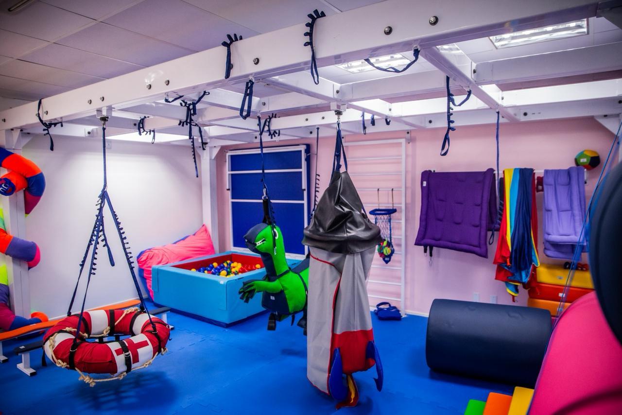 Sala de fisioterapia com brinquedos suspensos e piscina de bolinhas.
