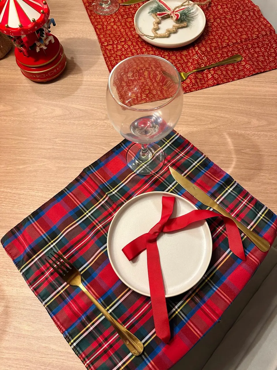 Foto em close-up de mesa posta com guardanapo xadrez sob prato branco, decorado com laço de fita vermelha, talheres dourados e taça de vidro.