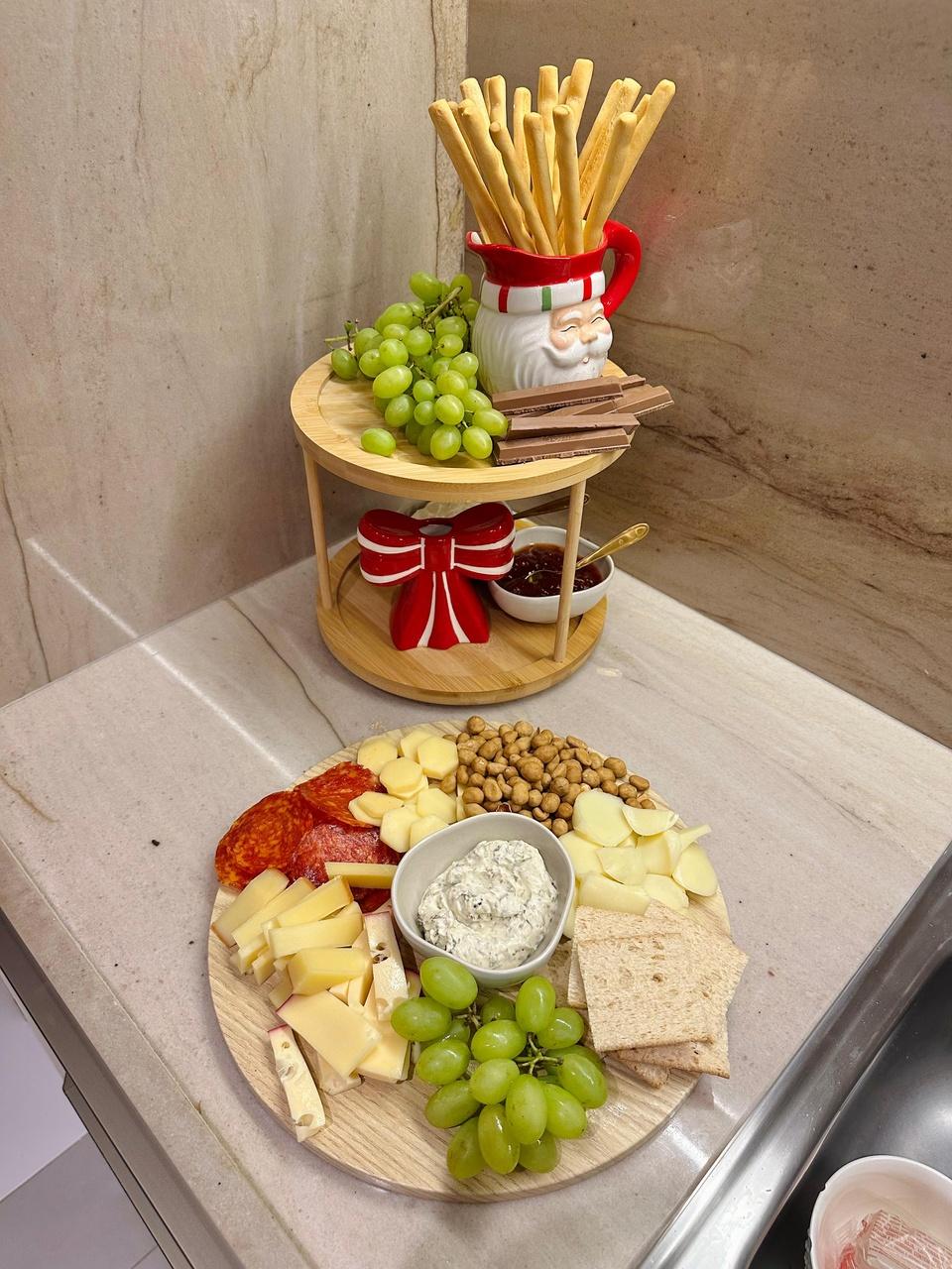 Tábua de frios e petiscos ao lado de um organizador giratório de dois andares com frutas, palitinhos em um pote de Papai Noel e decoração natalina.