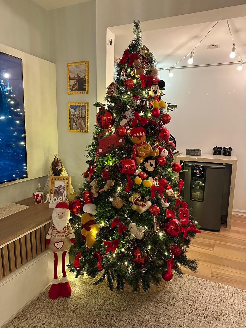 Árvore de Natal decorada com enfeites vermelhos, dourados e personagens, iluminada por luzes pisca-pisca, ao lado de um Papai Noel de pelúcia e móvel de madeira.