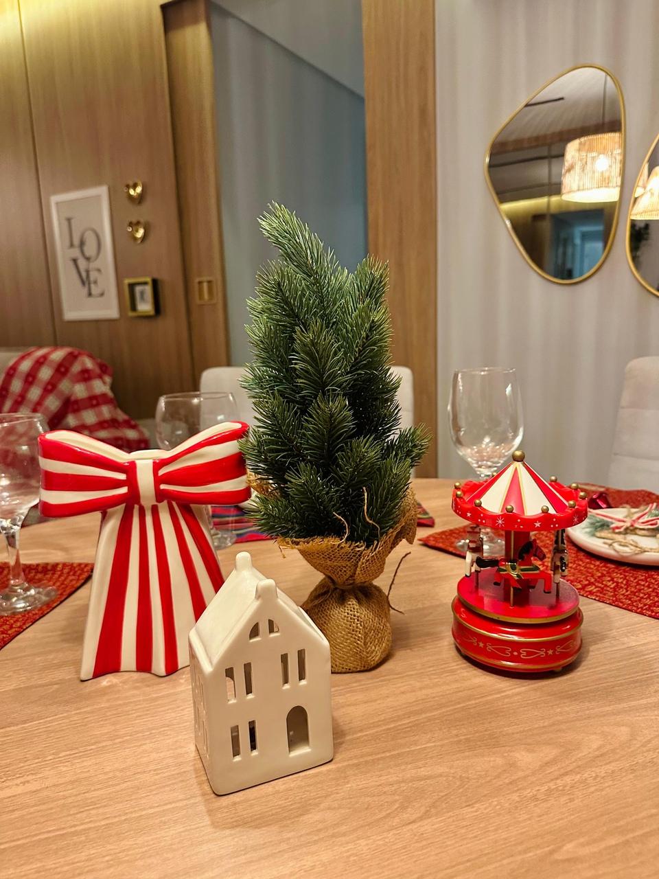 Centro de mesa natalino sobre mesa de madeira, composto por um pequeno pinheiro em base de juta, um laço listrado em vermelho e branco, uma casinha de cerâmica branca e um carrossel em miniatura. Os objetos possuem alturas variadas, criando um visual dinâmico.