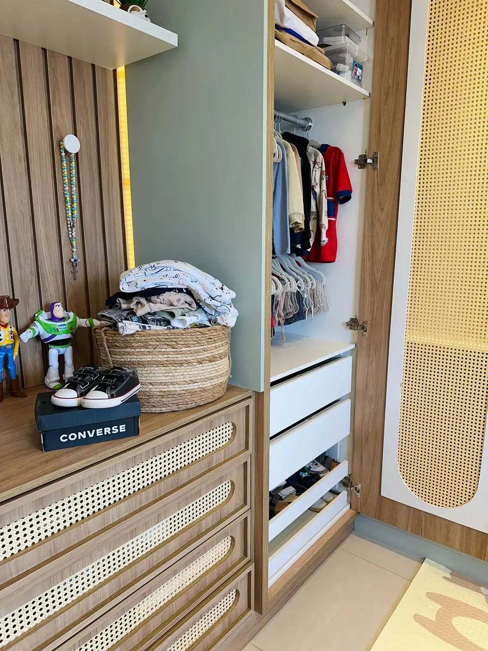 Um guarda-roupa infantil de madeira com detalhes em palhinha está com as portas abertas, revelando roupas penduradas e gavetas organizadas. Sobre a cômoda ao lado, há um cesto de palha cheio de roupas dobradas, um par de tênis em cima de uma caixa da Converse e bonecos do Woody e Buzz Lightyear.
