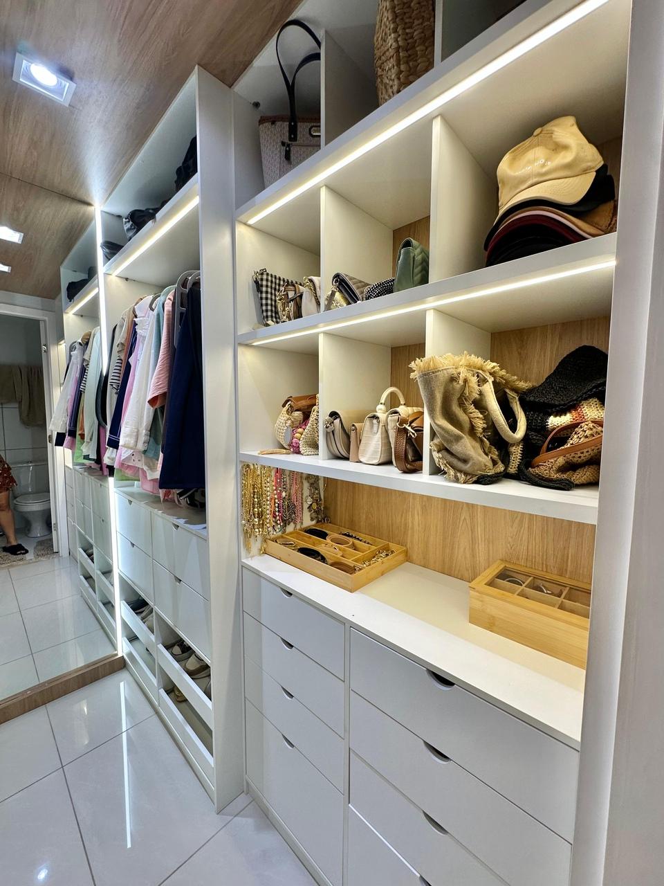 Um closet moderno e bem iluminado apresenta prateleiras brancas organizadas com bolsas, chapéus e acessórios, além de araras com roupas penduradas ao fundo. A parte inferior possui diversas gavetas brancas alinhadas, e uma bancada exibe organizadores de madeira para joias e óculos.