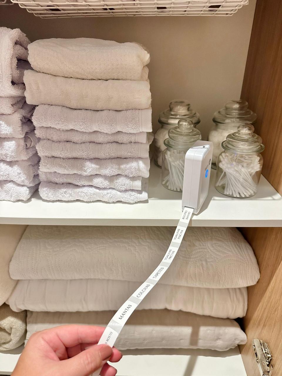 Uma prateleira de armário branca exibe toalhas dobradas, potes de vidro com algodão e cotonetes, além de uma rotuladora imprimindo etiquetas de identificação como 'mantas' e 'colchas'. Uma mão segura a fita de etiquetas recém-impressas, destacando o processo de organização e setorização do espaço.