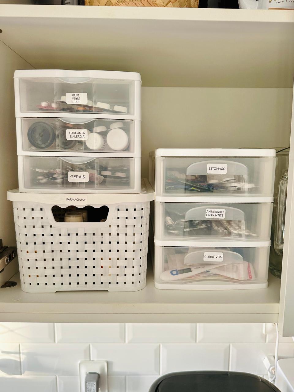 Uma prateleira branca organiza diversos medicamentos em pequenos gaveteiros de acrílico transparente e uma cesta plástica, todos devidamente identificados com etiquetas brancas. As etiquetas setorizam os itens por categorias, permitindo visualizar facilmente o conteúdo interno de cada compartimento.