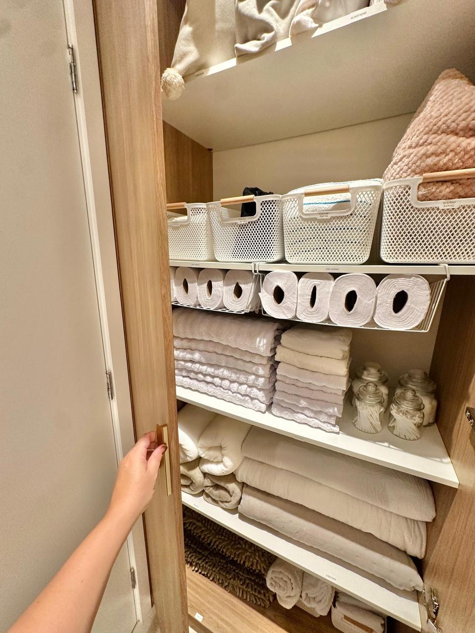 Uma mão abre a porta de madeira de um roupeiro perfeitamente organizado, revelando prateleiras brancas com cestos plásticos, toalhas dobradas em pilhas impecáveis e potes de vidro. O espaço utiliza suportes aramados para otimizar o armazenamento de rolos de papel, mantendo cada item setorizado e facilmente acessível.