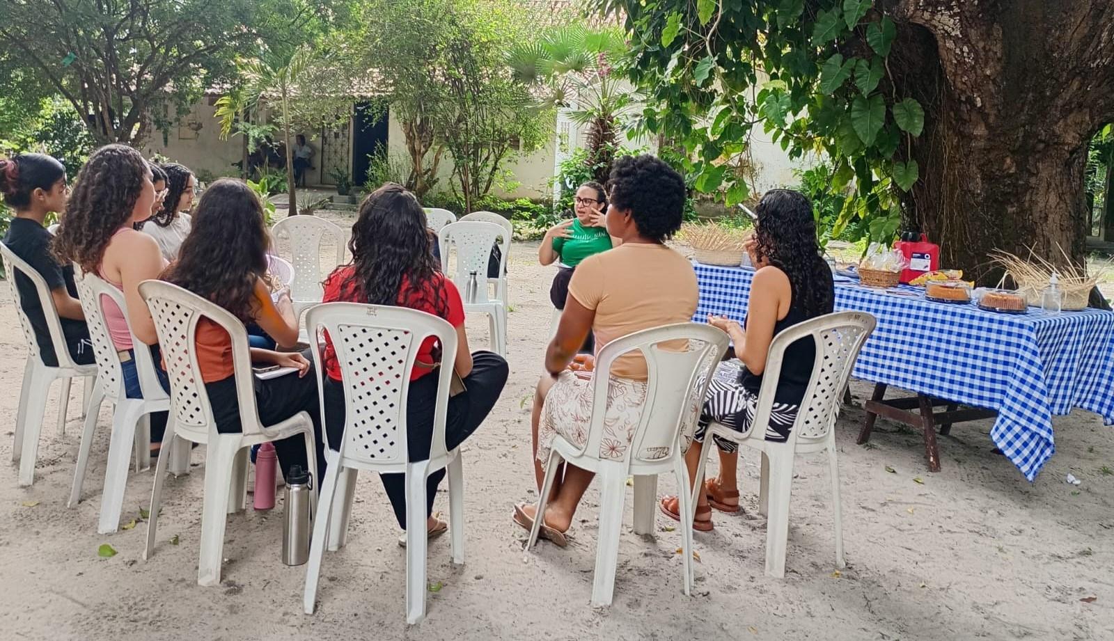 Grupo de pessoas participando de uma reunião ou oficina em sala de aula, com cadeiras dispostas em círculo e documentos na mão