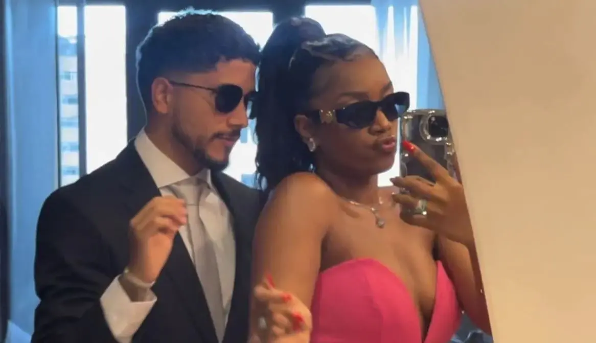 Duas pessoas posam para uma foto em frente a um espelho. Uma delas segura um celular para tirar a selfie e veste uma roupa elegante de cor rosa vibrante, com joias visíveis. A outra pessoa está ao lado, vestindo um traje formal escuro com camisa clara.