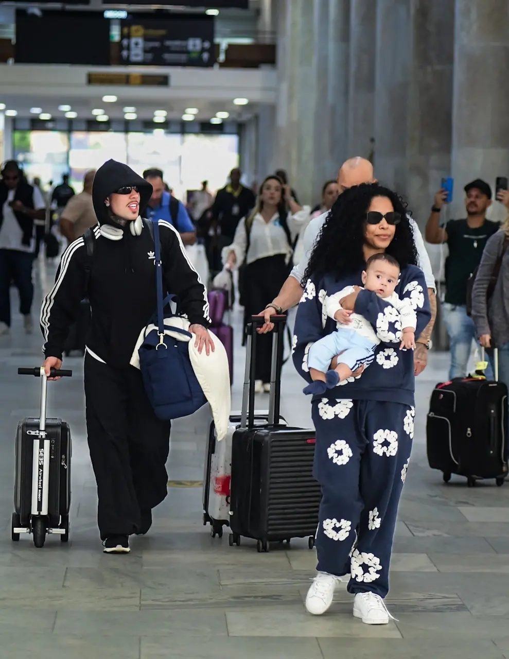 Duas pessoas caminham em um aeroporto movimentado, cada uma puxando uma mala de rodinhas. Uma delas veste um conjunto esportivo preto com listras brancas e carrega uma bolsa azul e um travesseiro branco. A outra pessoa usa um conjunto azul com estampa de flores brancas.