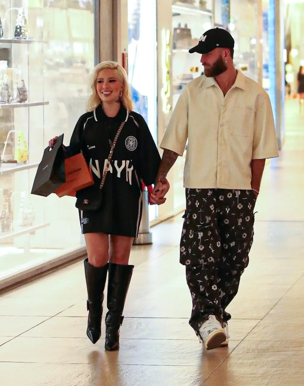Duas pessoas caminham de mãos dadas em um shopping. Uma delas veste uma camisa oversized preta com letras brancas, botas pretas de cano alto e carrega sacolas de compras. A outra pessoa usa uma camisa clara de manga curta, calças escuras com estampas e tênis brancos.