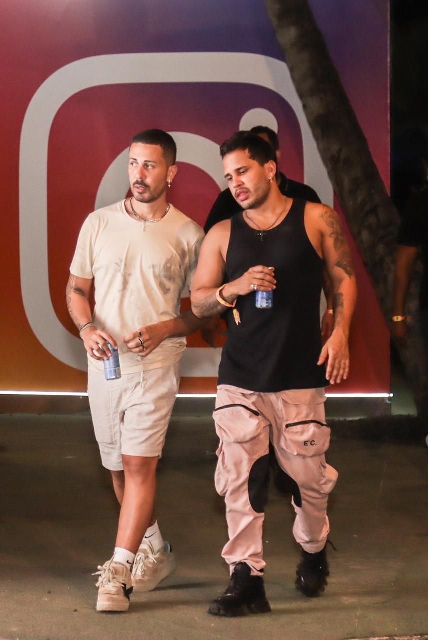 Duas pessoas caminham lado a lado em um espaço externo com piso de concreto, segurando latas de bebida. Uma delas veste camiseta clara e bermuda combinando, com tênis brancos, enquanto a outra usa regata preta e calça cargo rosa claro com bolsos grandes.