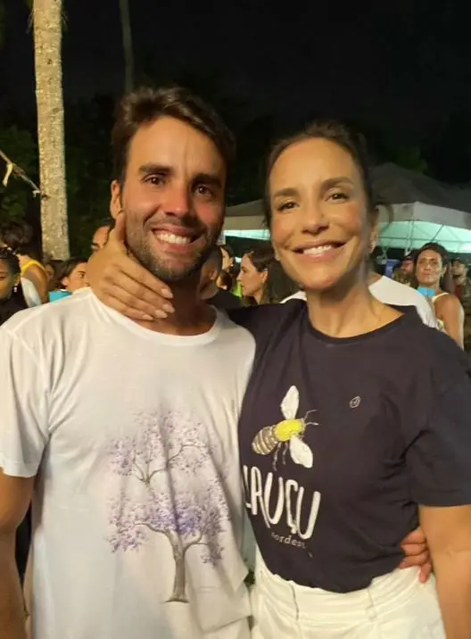 Uma mulher de camisa preta ao lado de um homem com blusa branca. Ela o abraça com a mão no pescoço e os dois estão sorrindo.