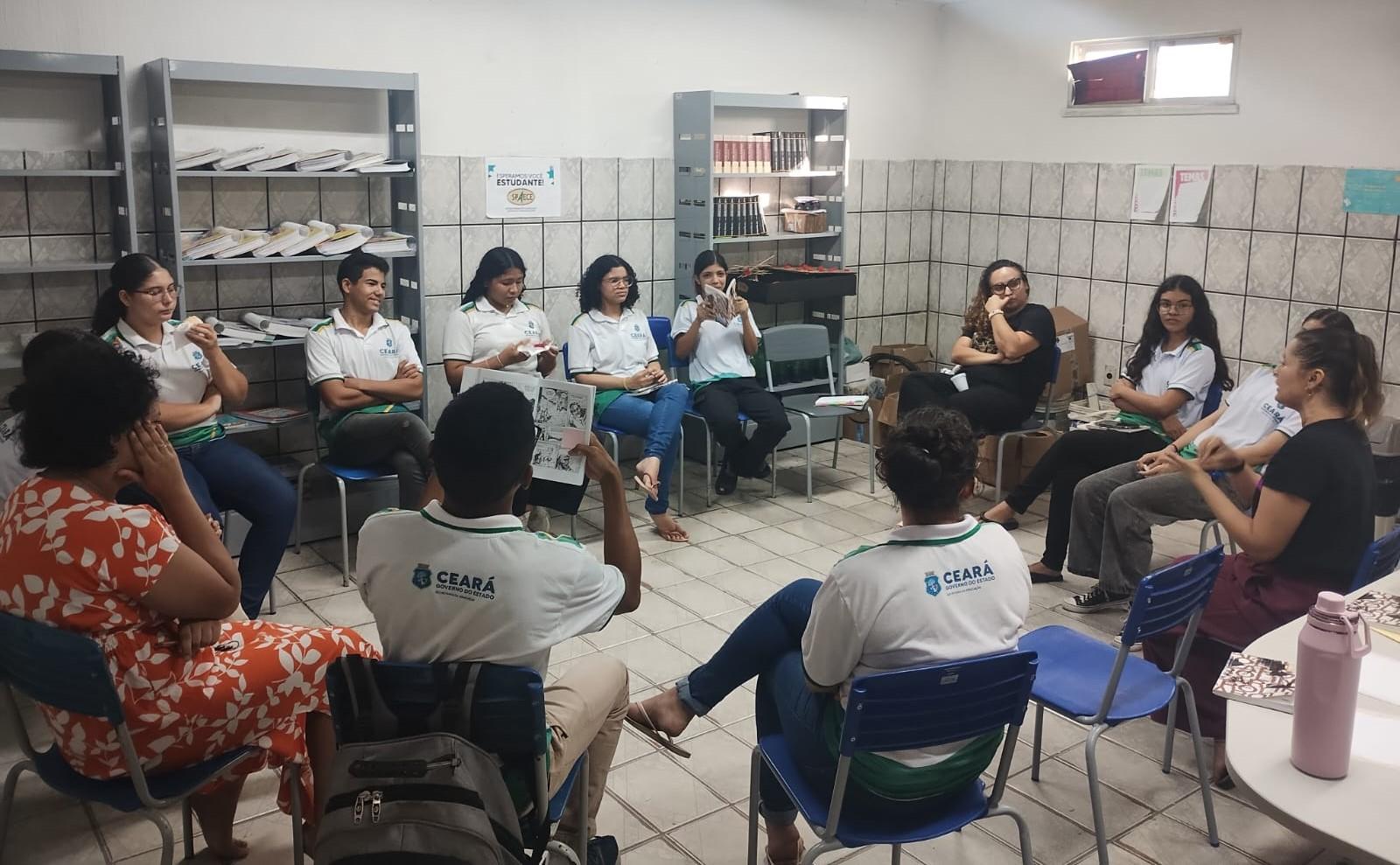Grupo de pessoas participando de uma reunião ou oficina em sala de aula, com cadeiras dispostas em círculo e documentos na mão