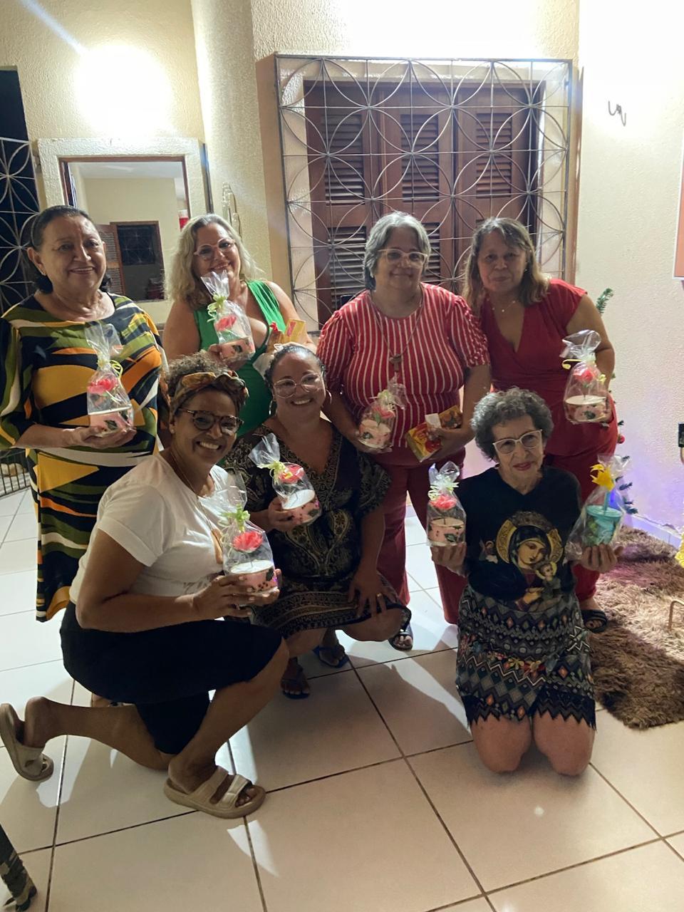 Na imagem, sete mulheres sorridentes posam juntas em um ambiente interno bem iluminado, celebrando uma confraternização. Quatro delas estão de pé ao fundo e três estão agachadas à frente. Quase todas seguram pequenos presentes embalados em celofane transparente com laços coloridos. Elas vestem roupas casuais e variadas, incluindo vestidos estampados, blusas coloridas e uma camiseta com a imagem de uma santa. O ambiente possui piso de cerâmica clara e, ao fundo, há uma porta de madeira protegida por uma grade de ferro trabalhada com desenhos circulares. À direita, nota-se parte de um tapete felpudo marrom e luzes decorativas.
