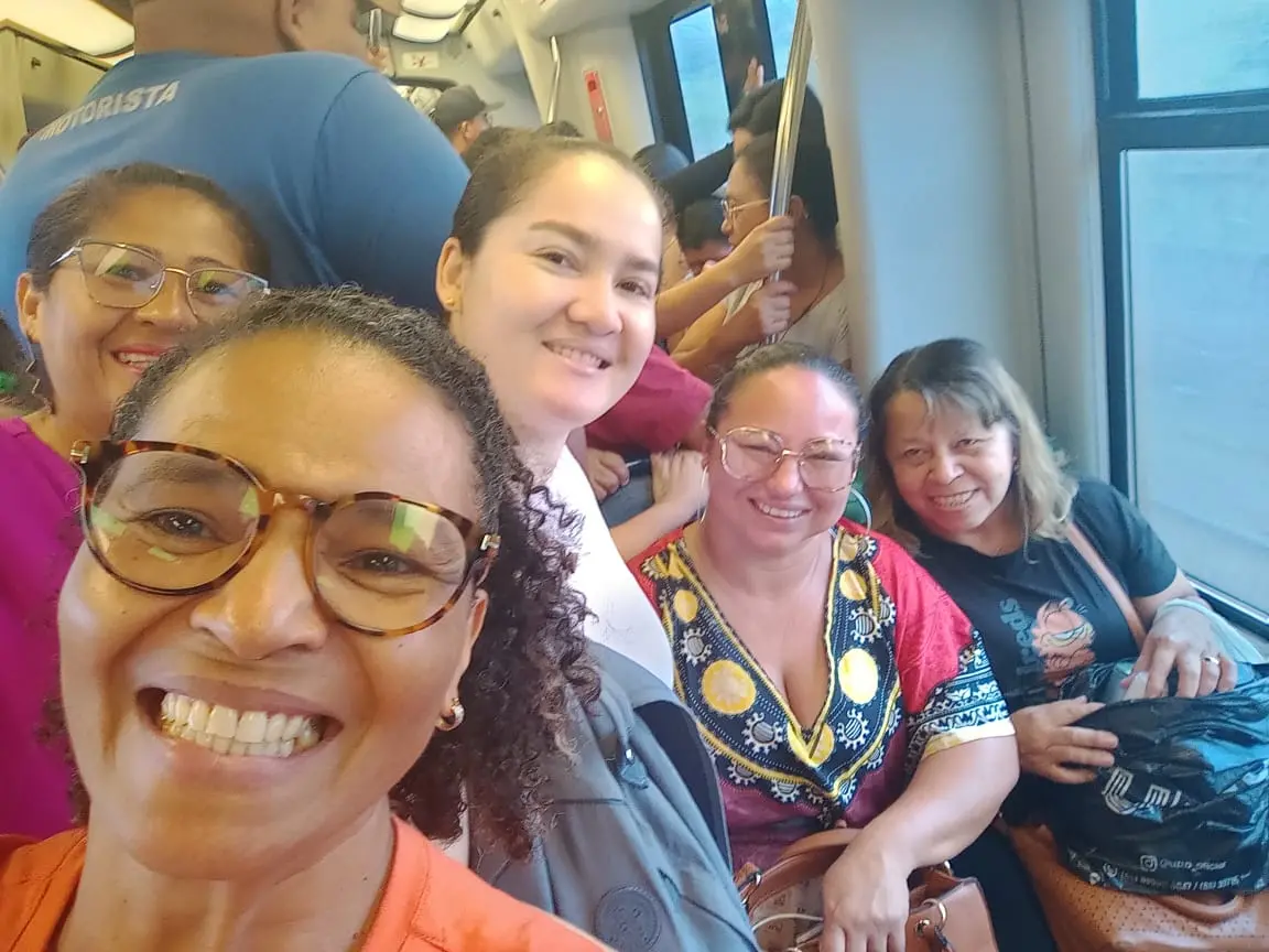 Na imagem, selfie em close de um grupo de cinco mulheres sorridentes dentro de um vagão de metrô. A mulher em primeiro plano, à esquerda, tem cabelos cacheados, usa óculos de armação tartaruga e exibe um sorriso largo. Ao lado e atrás dela, as outras amigas aparecem em diferentes planos, ocupando os assentos e o corredor do trem. Elas vestem roupas coloridas e casuais. Ao fundo, é possível ver outros passageiros e as janelas do metrô, indicando que o transporte está em movimento durante o dia.
