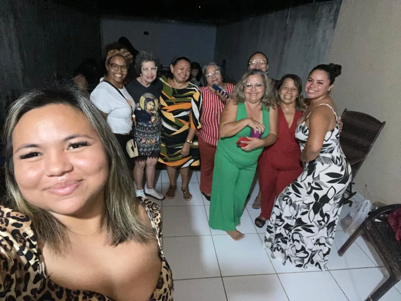 Na imagem, selfie tirada por uma jovem em primeiro plano com estampa de oncinha, mostrando um grupo de nove mulheres reunidas em uma sala com piso claro. As amigas ao fundo estão em pé, sorrindo e abraçadas, vestindo roupas variadas como macacões coloridos, vestidos estampados e camisetas casuais. O ambiente é simples, com paredes claras e algumas cadeiras de vime visíveis ao lado.