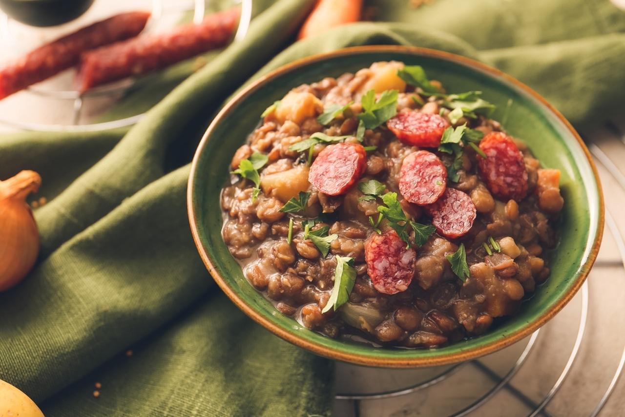 Prato fundo verde com ensopado de lentilhas, decorado com fatias de linguiça e folhas de salsa fresca; ao fundo, aparecem ingredientes como linguiça e cebola sobre um pano verde.