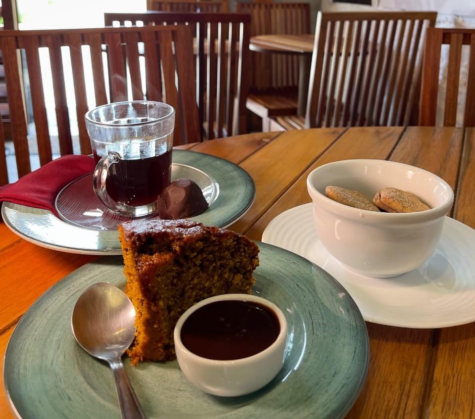 Imagem de uma mesa de café da manhã com bolo de cenoura, chocolate, suco de uva, biscoitos e utensílios de porcelana em ambiente aconchegante.