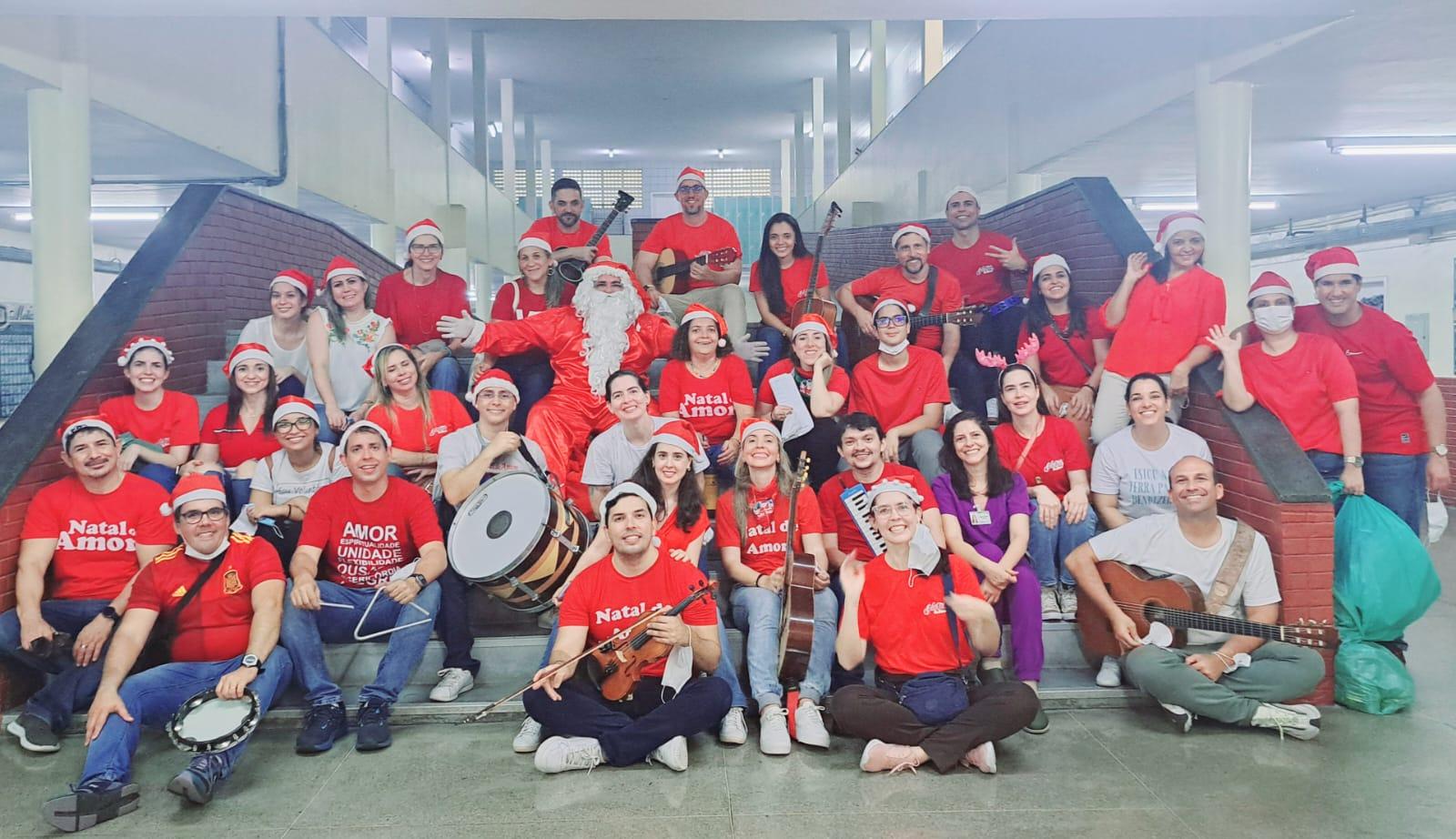 Na imagem, uma fotografia em plano aberto mostra um grande grupo de voluntários reunidos em uma escadaria interna de um prédio. Quase todos vestem camisetas vermelhas — algumas com a frase 