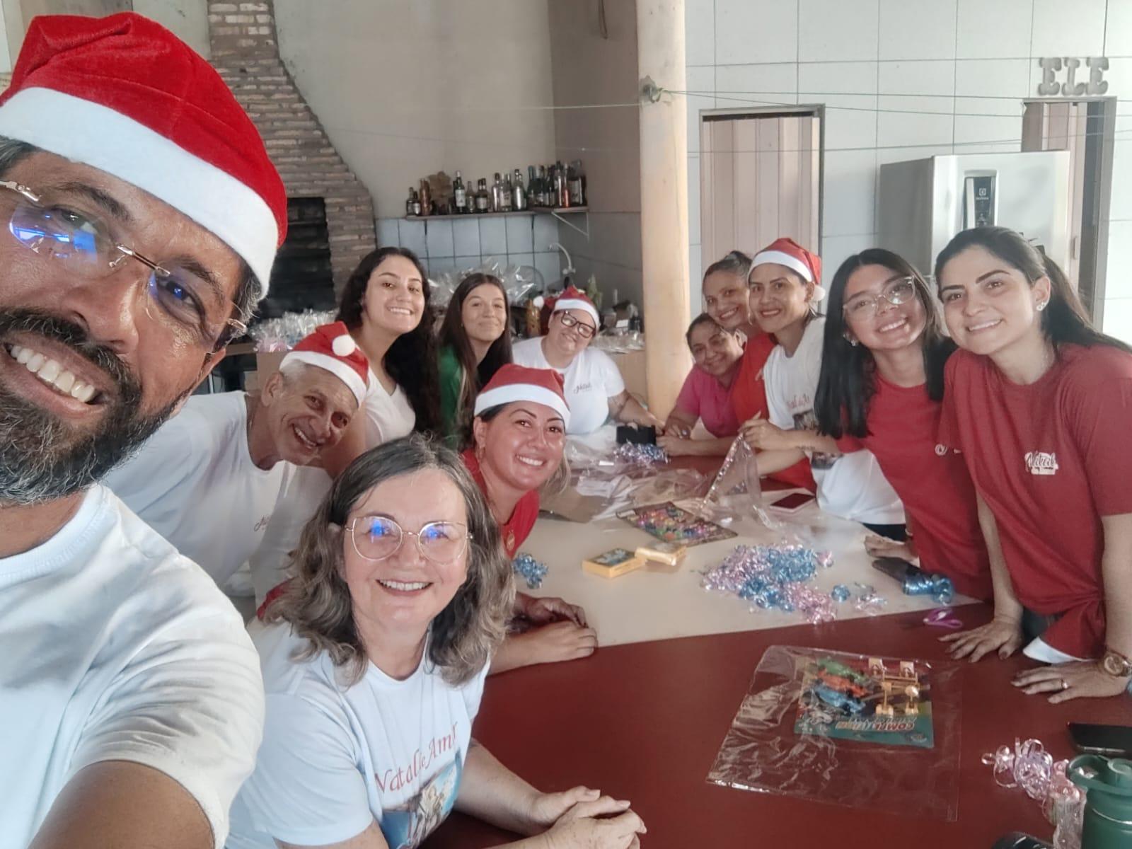 Na imagem, uma fotografia em plano médio mostra um grupo de pessoas em uma área coberta organizando doações de roupas infantis e fraldas. No centro, mesas estão repletas de pilhas organizadas de roupas coloridas (tons de amarelo, azul e rosa) e pacotes de fraldas descartáveis ao fundo. À esquerda, uma mulher de óculos e camiseta branca observa o trabalho, enquanto à direita, um homem de camiseta verde e uma mulher organizam e embalam as peças de roupa em sacos plásticos transparentes. No canto inferior esquerdo, há um grande saco plástico verde cheio de donativos. O ambiente é amplo, com colunas claras e telhado aparente, sugerindo um esforço comunitário de voluntariado e solidariedade.