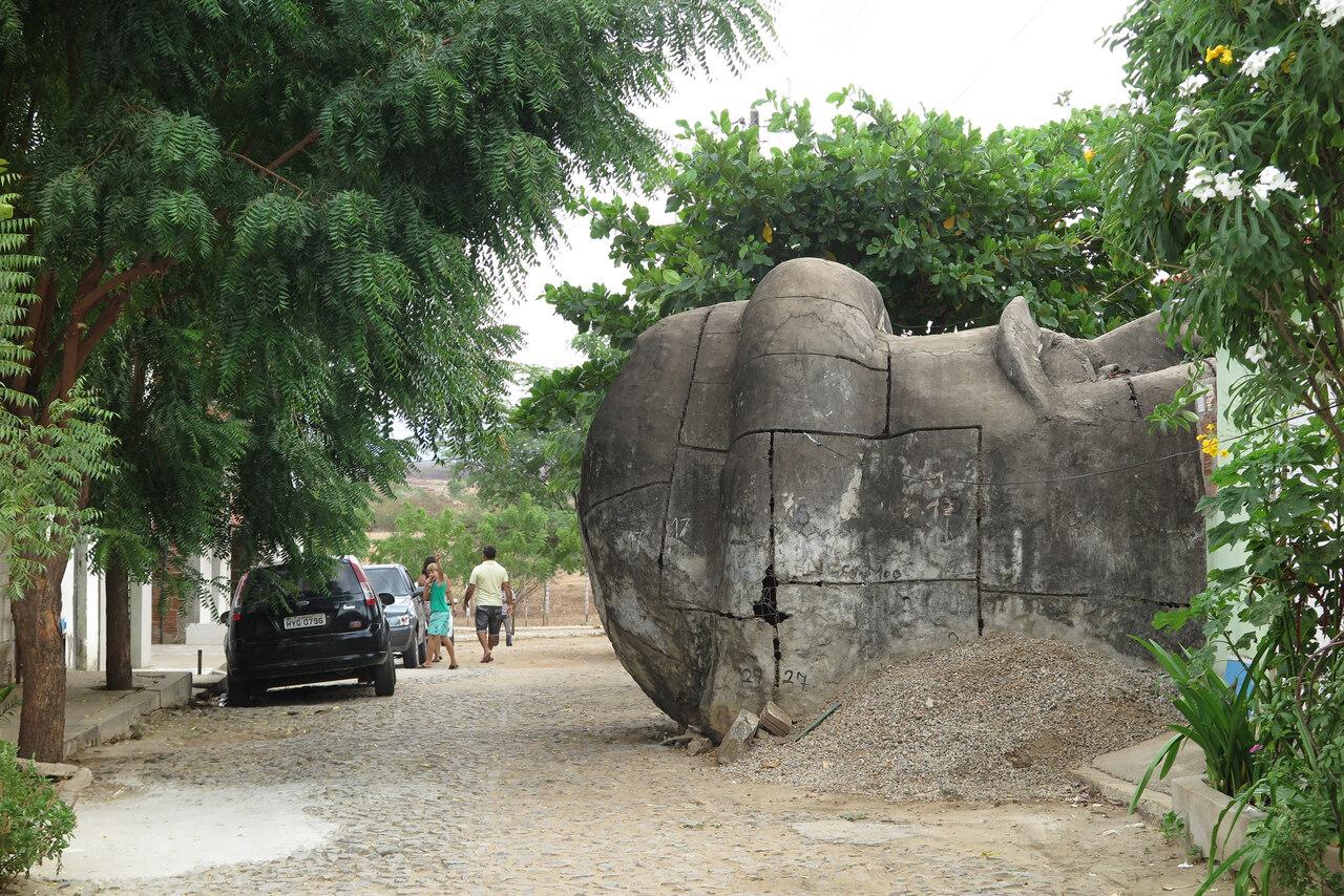 Uma cabeça colossal de concreto da estátua de Santo Antônio repousa de lado em uma rua de paralelepípedos, cercada por árvores verdes e casas simples. A estrutura monumental apresenta marcas de montagem e numeração, contrastando com o movimento cotidiano de pedestres e carros ao fundo sob o céu nublado.