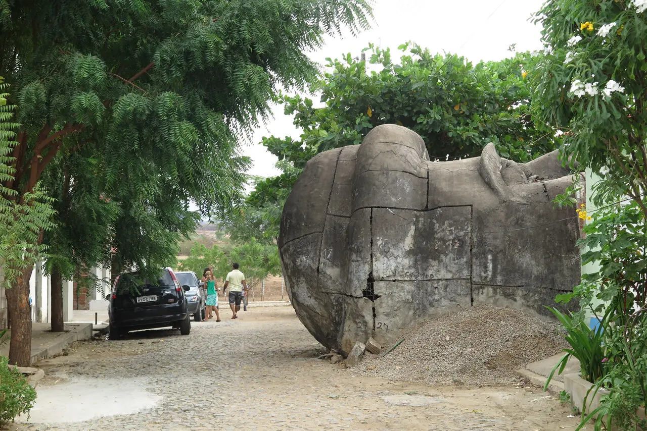 Uma cabeça colossal de concreto da estátua de Santo Antônio repousa de lado em uma rua de paralelepípedos, cercada por árvores verdes e casas simples. A estrutura monumental apresenta marcas de montagem e numeração, contrastando com o movimento cotidiano de pedestres e carros ao fundo sob o céu nublado.