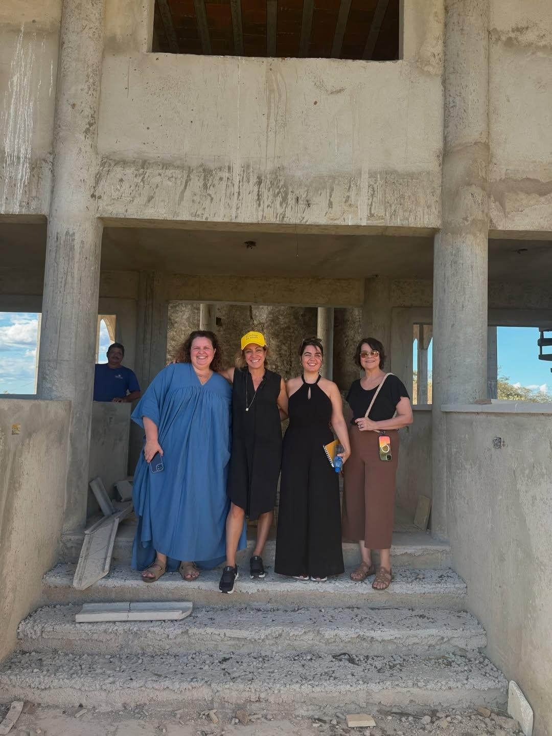 Quatro mulheres sorriem posicionadas em uma escadaria de concreto dentro de uma construção inacabada durante uma visita de locação em Caridade, Ceará. A estrutura bruta de cimento enquadra o grupo, que veste roupas casuais.