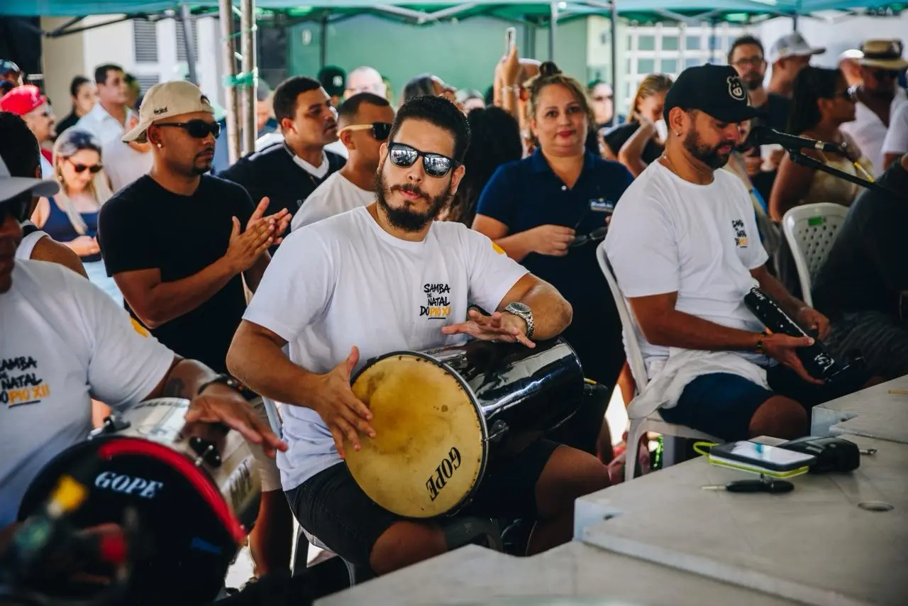 Grupo de pessoas participa de uma roda de samba ao ar livre. Em primeiro plano, um homem com barba, óculos escuros e camiseta branca toca um tantan sentado, enquanto outros músicos ao redor batem palmas e tocam instrumentos de percussão. Ao fundo, o público acompanha a apresentação sob tendas verdes, em um ambiente de confraternização e música.