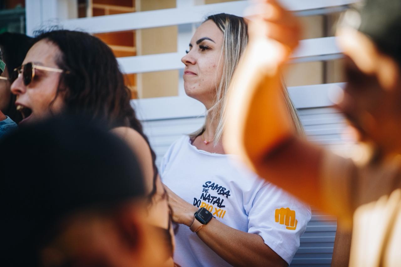 Mulher em pé acompanha uma roda de samba, vestindo camiseta branca com a inscrição “Samba de Natal do Pio XII”. Ela observa a apresentação com expressão atenta, enquanto ao redor aparecem outras pessoas, algumas desfocadas, batendo palmas e cantando em um ambiente festivo.