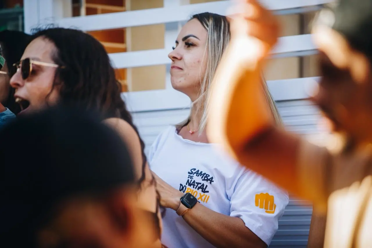 Mulher em pé acompanha uma roda de samba, vestindo camiseta branca com a inscrição “Samba de Natal do Pio XII”. Ela observa a apresentação com expressão atenta, enquanto ao redor aparecem outras pessoas, algumas desfocadas, batendo palmas e cantando em um ambiente festivo.