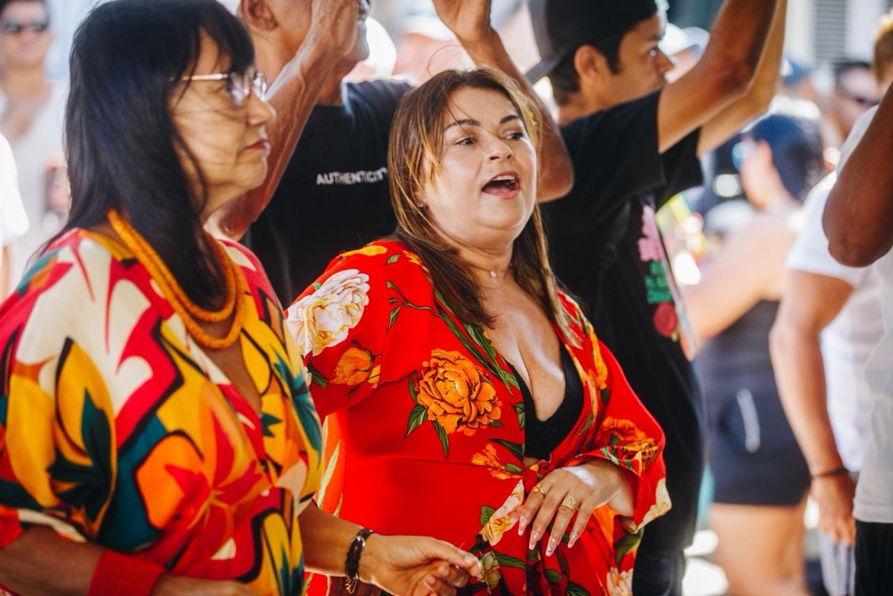Duas mulheres participam animadas de uma roda de samba. Em primeiro plano, uma delas usa vestido vermelho com estampa floral e canta com expressão alegre; ao lado, outra mulher veste roupa colorida e observa a apresentação. Ao fundo, outras pessoas aparecem com os braços erguidos, batendo palmas e acompanhando a música em um ambiente festivo.