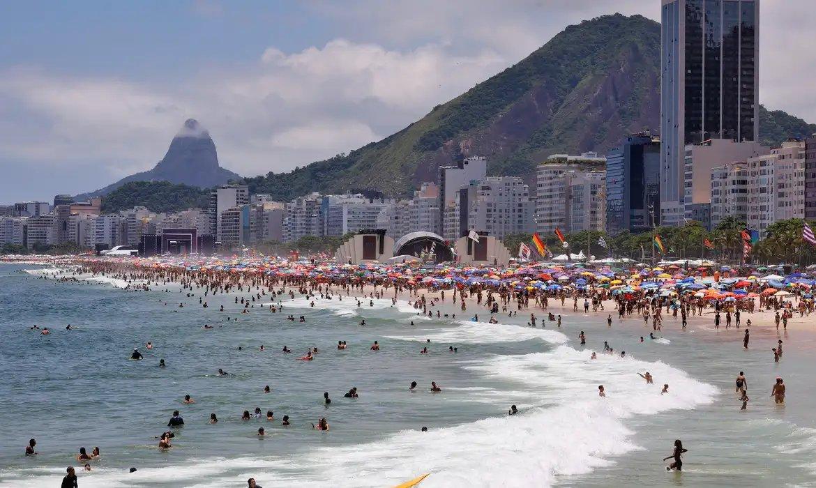 Vista panorâmica de uma praia do Rio de Janeiro lotada, com pessoas no mar e na areia sob guarda-sóis coloridos.
