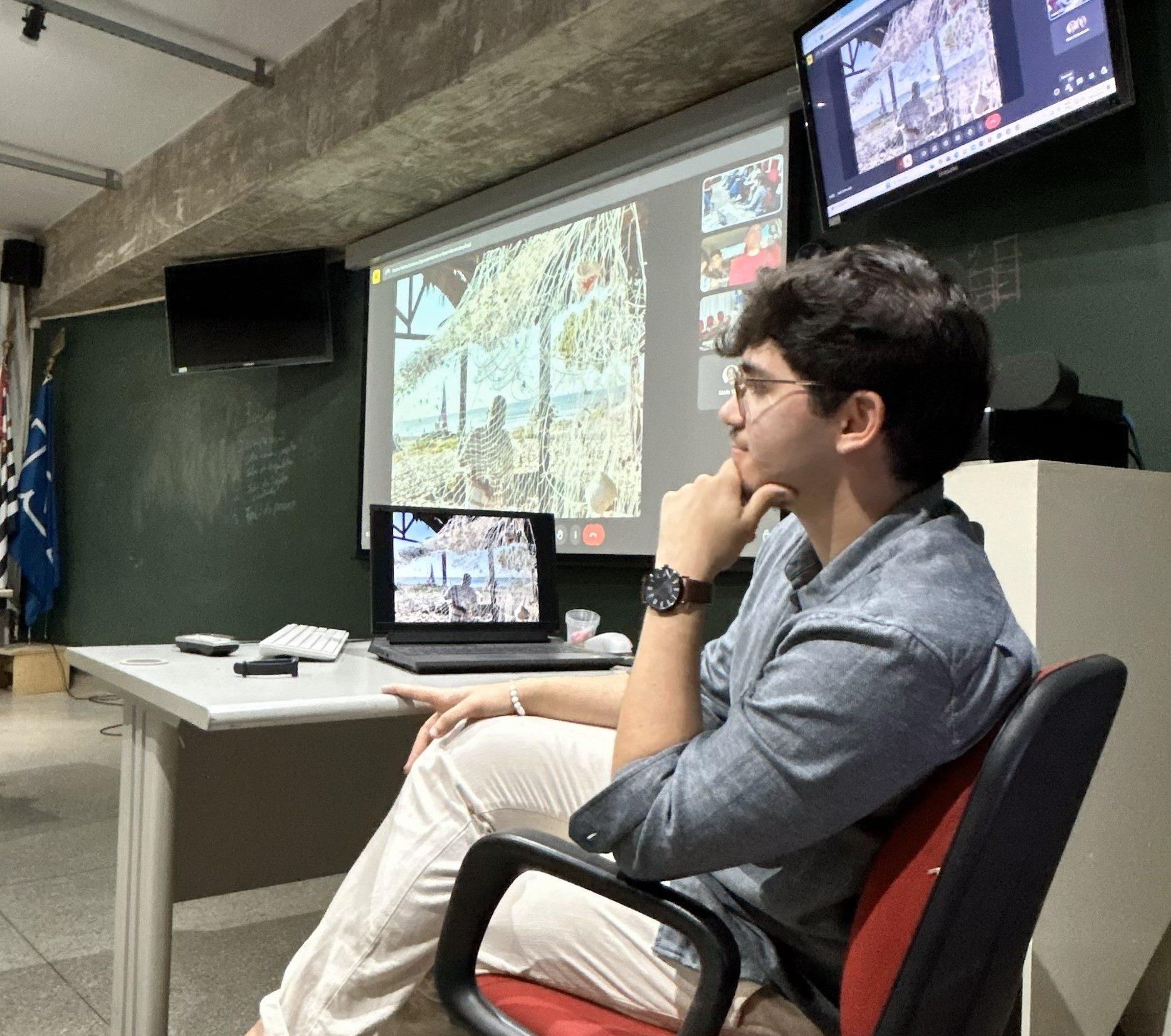 Pessoa assistindo a uma apresentação ou vídeo sobre naturezas variadas em uma sala de reuniões ou sala de aula, com tela grande e laptop.