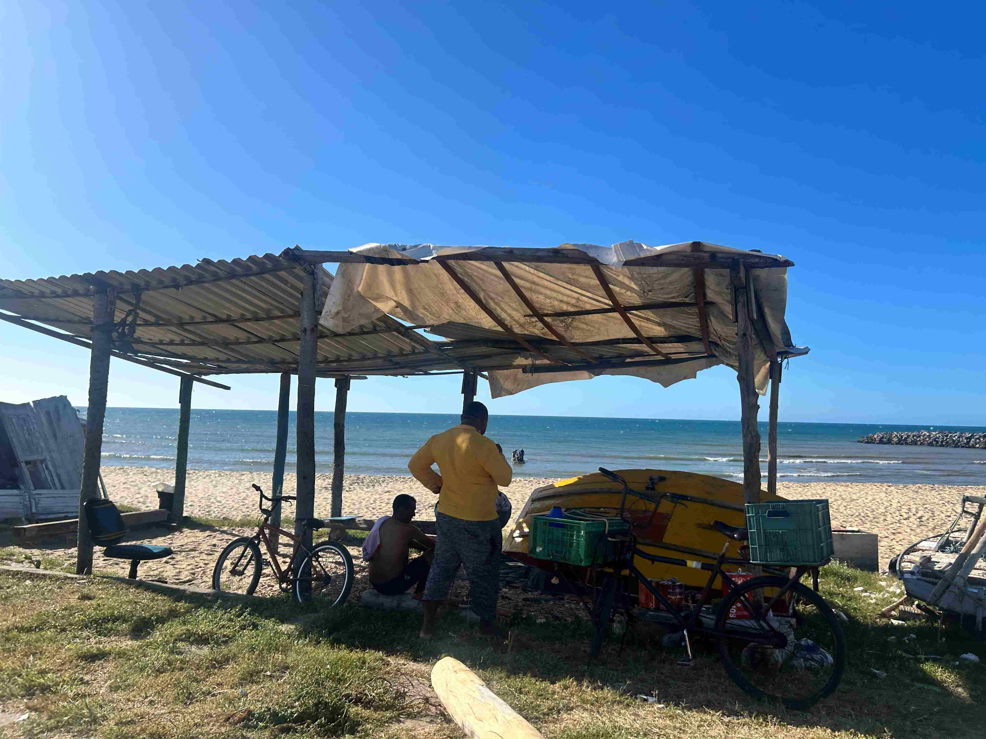 Imagem de uma cabana simples na praia com duas pessoas próximas, uma pedalando uma bicicleta e uma prancha de surf ao lado sob céu azul, no Pirambu, em Fortaleza.