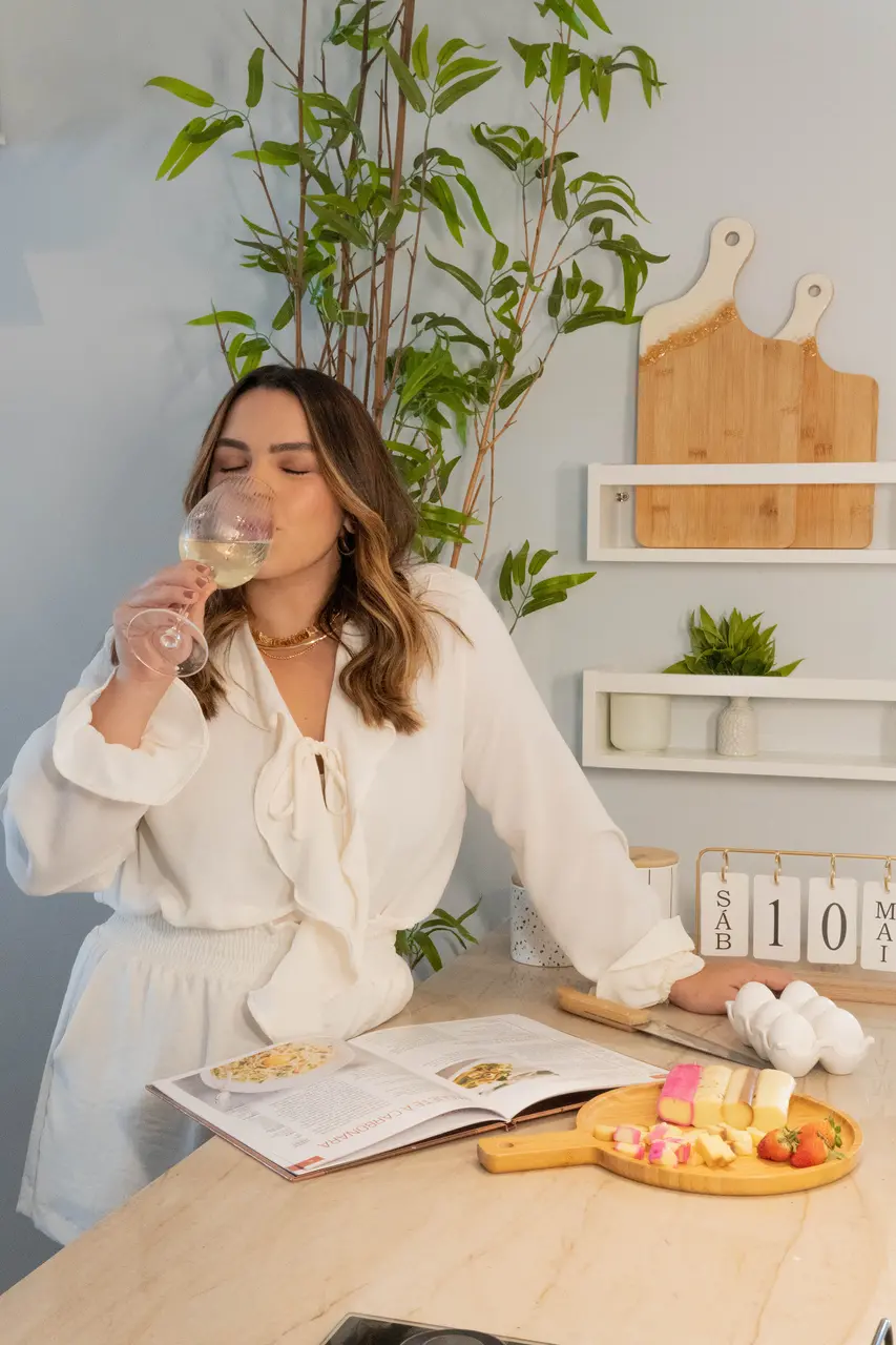 Mulher vestida de branco bebendo uma taça de vinho verde em um cenário de casa, com prateleiras de utensilios de cozinha.