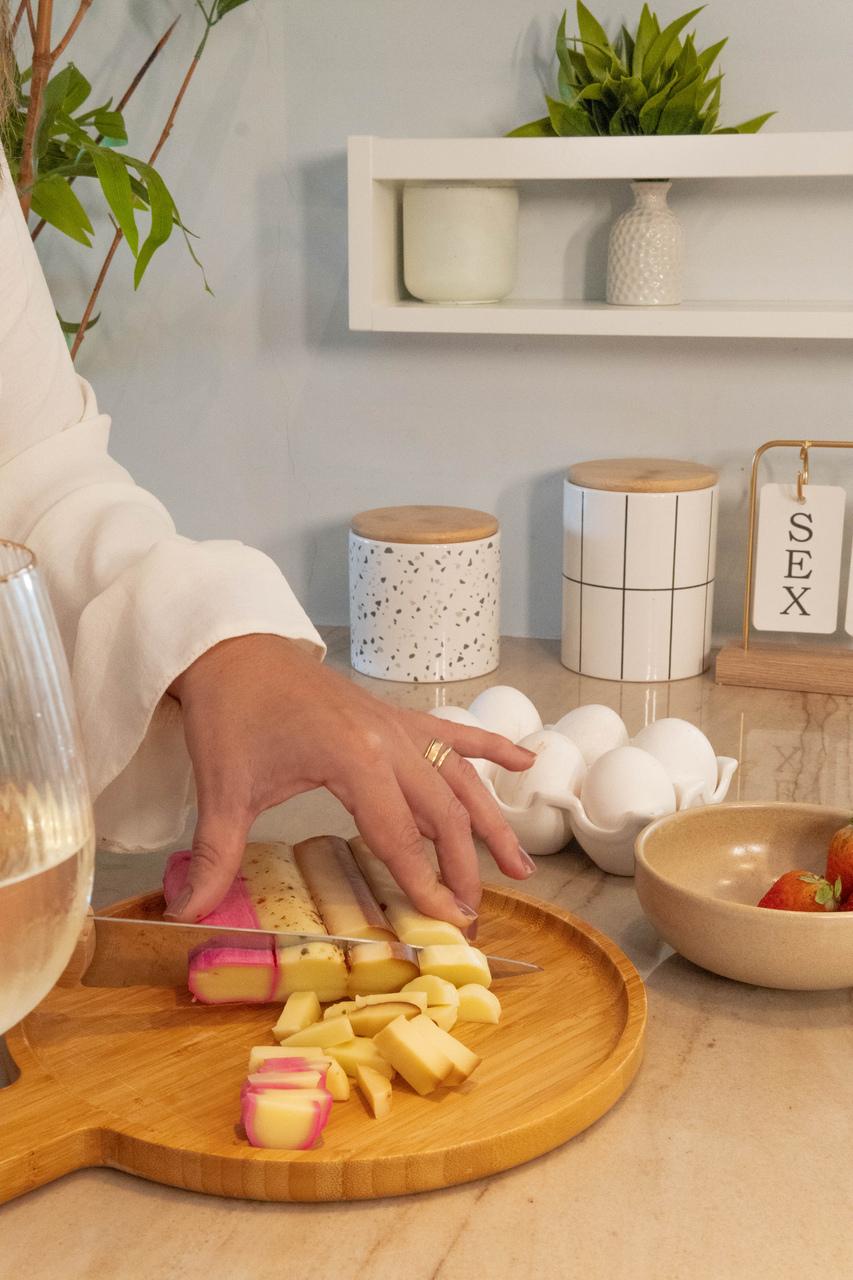 Mão de mulher em destaque cortando quatro tipos de queijo em uma tábua de madeira.