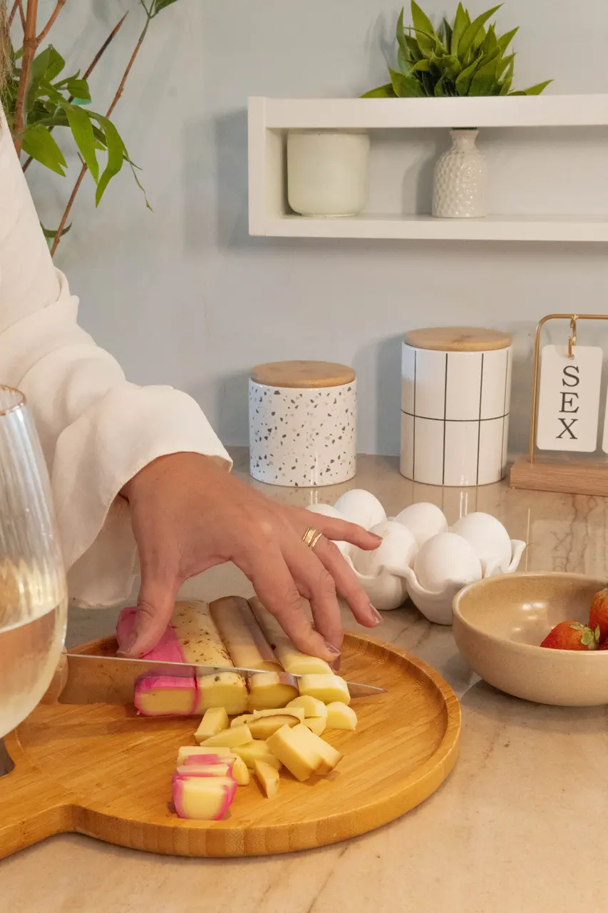 Mão de mulher em destaque cortando quatro tipos de queijo em uma tábua de madeira.